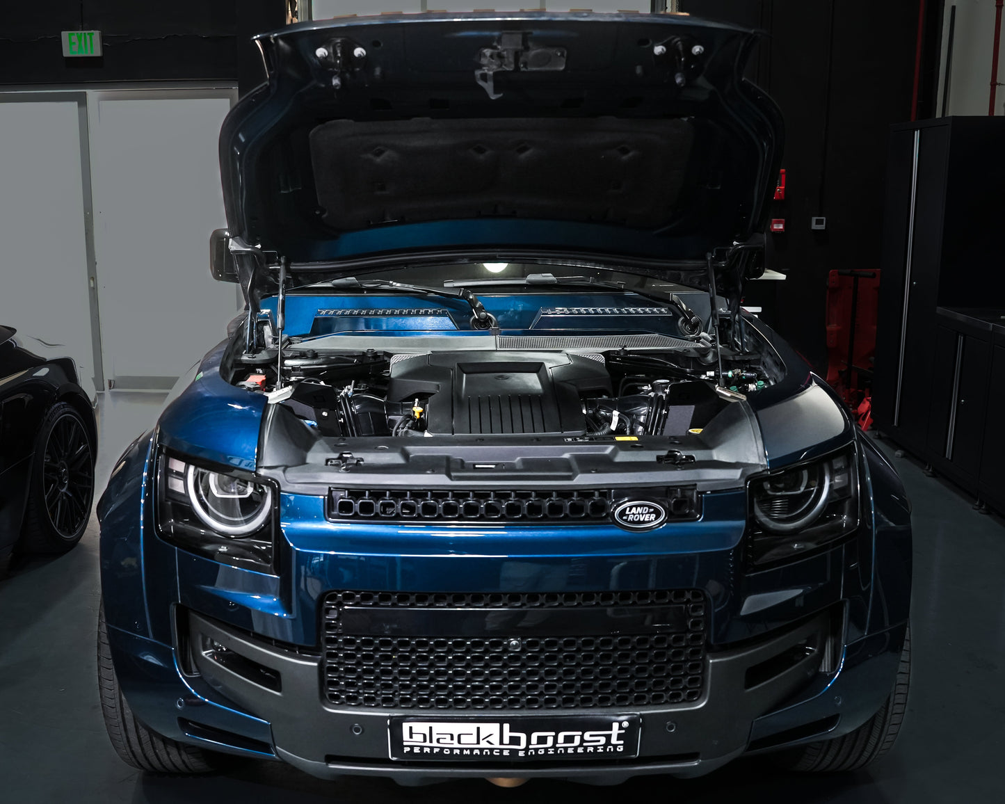 A blue metallic Land Rover with its hood open, showing a custom engine bay with a black and blue Lancer engine. The vehicle is parked in a dark garage setting with a custom grille and a 'Blackboost' license plate frame.