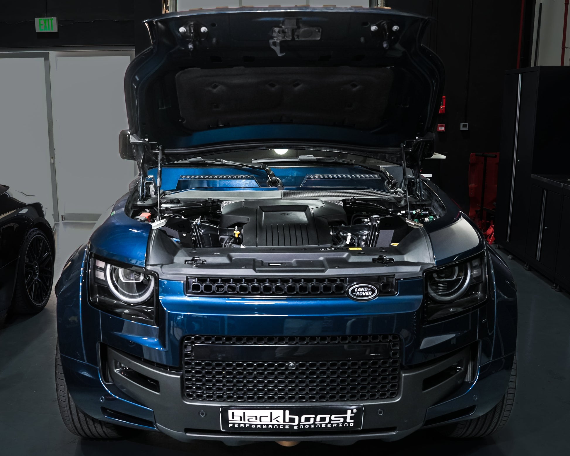 A blue metallic Land Rover with its hood open, showing a custom engine bay with a black and blue Lancer engine. The vehicle is parked in a dark garage setting with a custom grille and a 'Blackboost' license plate frame.