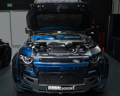A blue metallic Land Rover with its hood open, showing a custom engine bay with a black and blue Lancer engine. The vehicle is parked in a dark garage setting with a custom grille and a 'Blackboost' license plate frame.